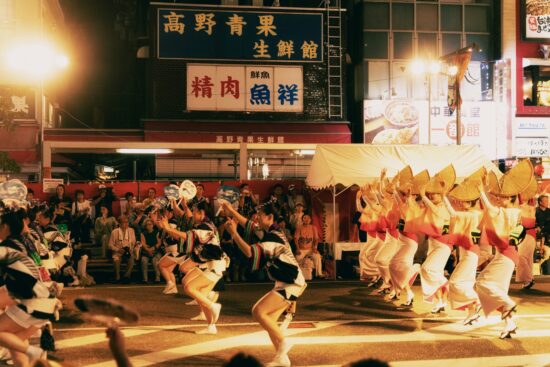 Awa Odori 2025 dancers performing in white yukata costumes with yellow festival lanterns on a Tokushima street at night, with spectators watching from storefronts